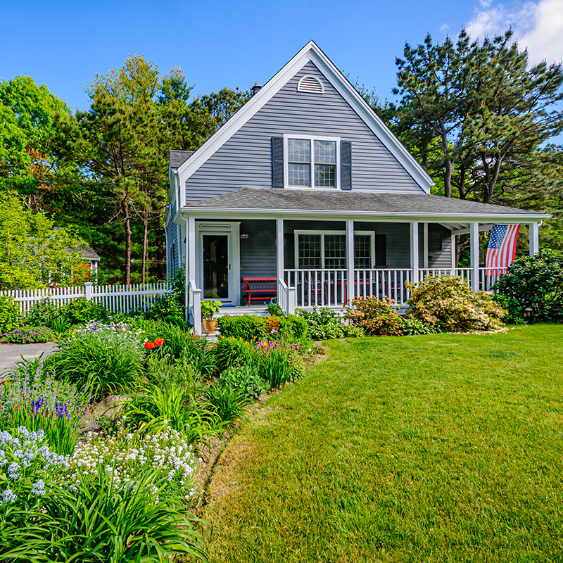 Springtime gardens along the driveway of a small single family home on Cape Cod with a flag flying from the front porch, and the surrounding grounds